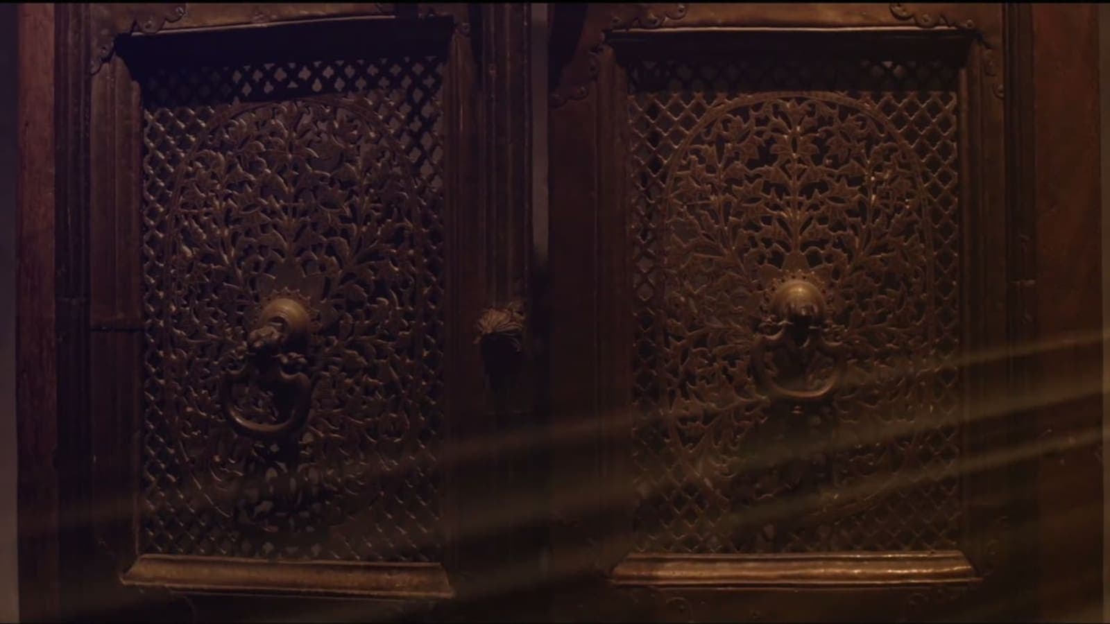 A still from the museum’s teaser film: carved wooden doors with pierced brass panels and ring handles, lit by shafts of light.